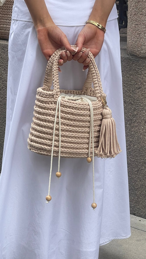 Kvinna i vit klänning håller en beige handvirkad Bucket-väska med flätat handtag och dragsko.