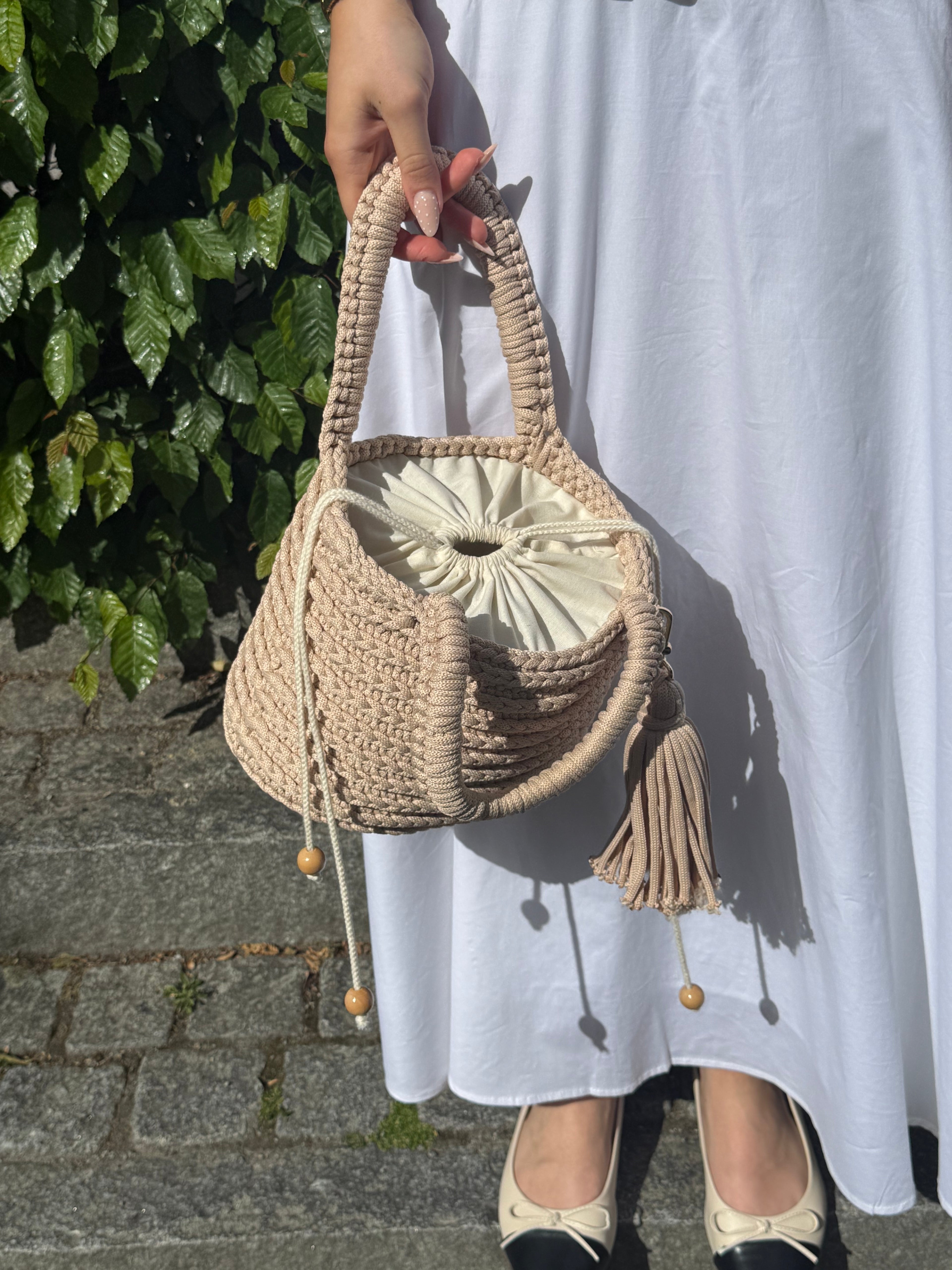 Beige handvirkad Bucket-väska buren till vit klänning, visad från sidan med dragskostängning och tofs.